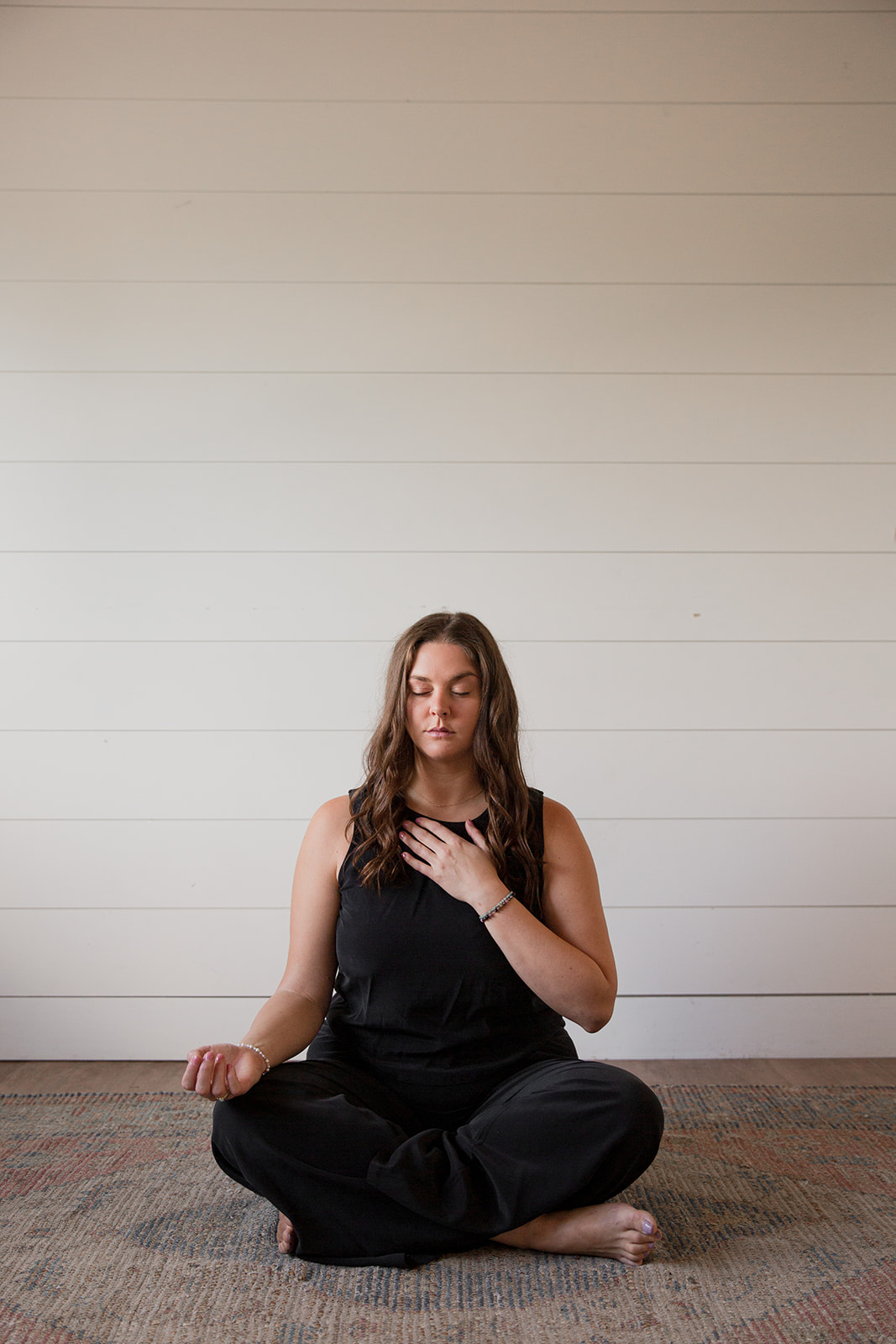 Heather Flynn seated cross-legged in meditation with a hand on her heart against a white shiplap wall